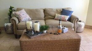 A living room with a beige sofa, two patterned throw pillows, a woven coffee table with candles, a small plant, and decorative items.