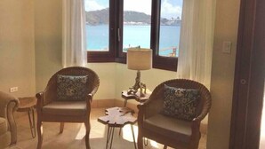 A corner room with two wicker chairs, a small table, and a lamp. A large window offers a view of the sea and hills.