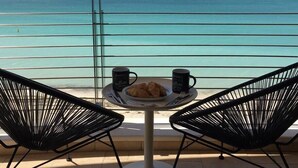 A table with two black chairs, a plate with croissants, and two black mugs on a balcony overlooking a turquoise sea.