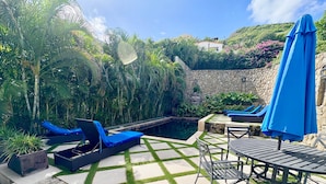 Ein Poolbereich mit Liegestühlen und einem Tisch, umgeben von üppiger Bepflanzung und einer Steinmauer.