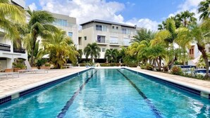 A rectangular swimming pool with clear blue water, surrounded by palm trees and a multi-story building with balconies. - Simpson bay 3bdr / 3,5 bath with pools - GENERATOR (Simpson Bay)