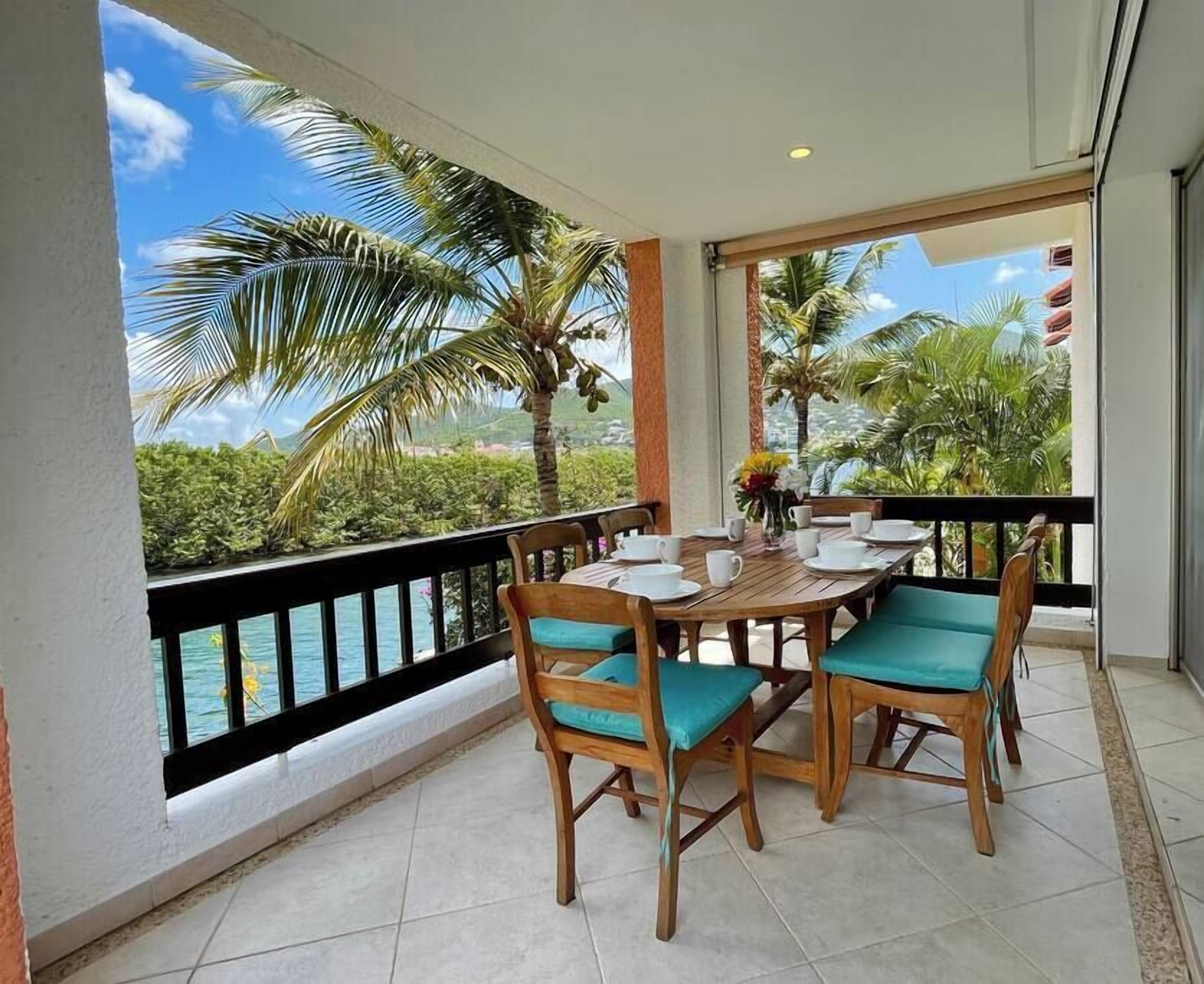 A balcony with a wooden table and chairs, a view of the ocean and palm trees, and a tiled floor.