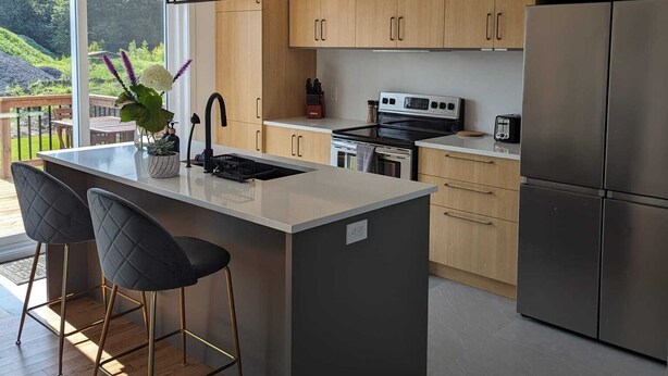 Une cuisine moderne avec des meubles en bois, un îlot central doté de tabourets de bar, des appareils électroménagers en acier inoxydable et une vue sur un paysage verdoyant grâce à de grandes fenêtres.
