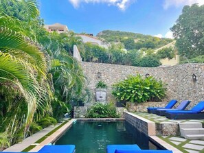 Un espace piscine agrémenté d’un mur en pierre, de palmiers et de chaises longues.