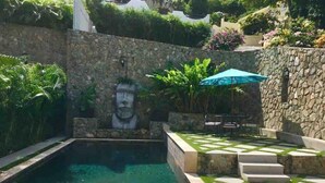 Une piscine bordée d’un mur en pierre et ornée d’une fresque, entourée de verdure et d’une terrasse carrelée.