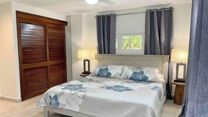 Une chambre à coucher avec un lit, deux lampes de chevet, une fenêtre avec des rideaux, un ventilateur de plafond et une porte en bois avec des stores.