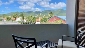 Un balcon avec deux chaises et une table, donnant sur un quartier résidentiel avec des maisons et des palmiers, sous un ciel partiellement nuageux.