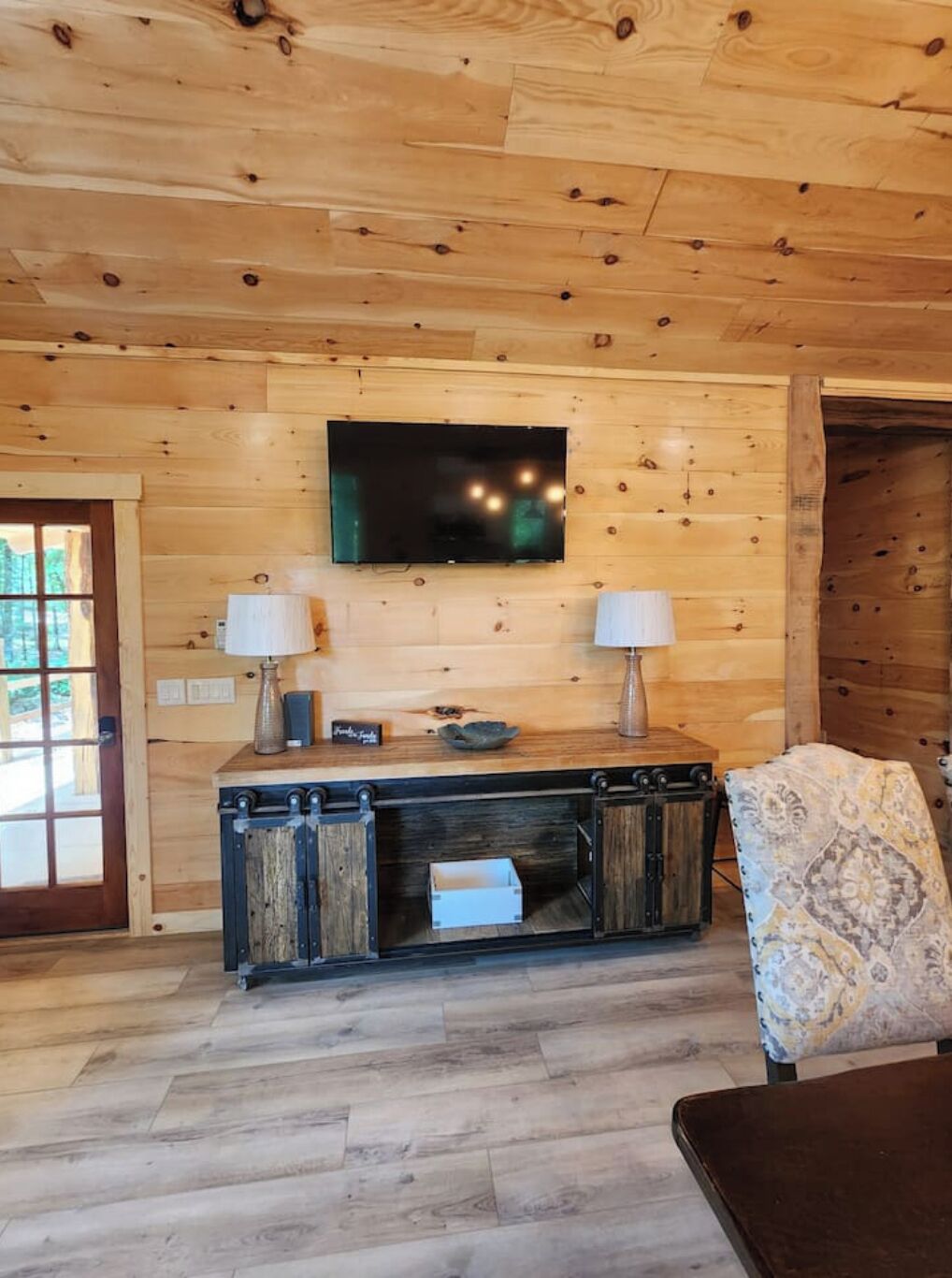 A wooden interior with a TV mounted on the wall, a wooden cabinet with two lamps, and a patterned chair.