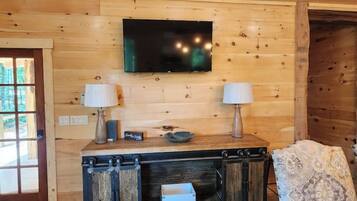 A wooden interior with a TV mounted on the wall, a wooden cabinet with two lamps, and a patterned chair.