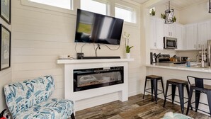 A modern living room with a fireplace, a television mounted above it, and a kitchen area with white cabinets and a bar stool.