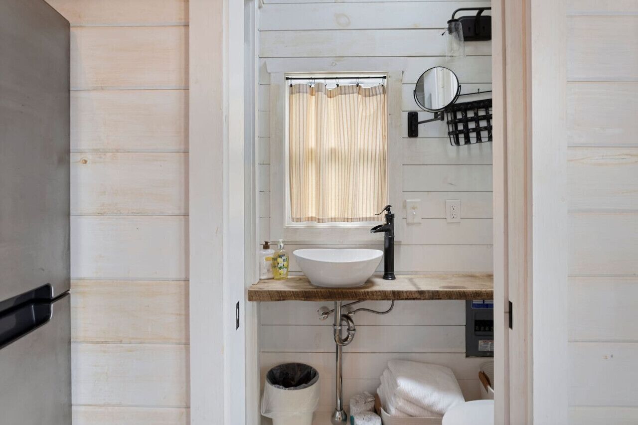 A compact bathroom with a wooden vanity, a white sink, a mirror, and a window with a striped curtain.