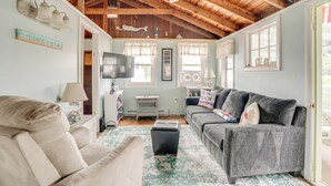 Un salon avec un plafond haut, des poutres en bois, un ventilateur de plafond, un canapé beige, un canapé gris, une table basse et un téléviseur posé sur un meuble.