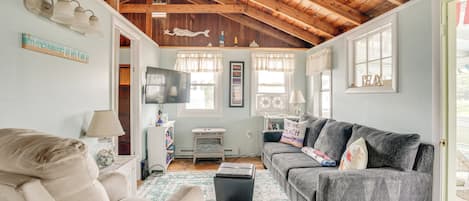 A living room with a high ceiling, wooden beams, a ceiling fan, a beige sofa, a gray sofa, a coffee table, and a TV on a stand.