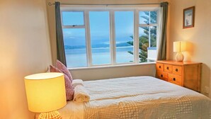 Ein ordentlich bezogenes Bett mit einem gemusterten Überwurf, einer Nachttischlampe und Blick auf das Meer durch ein großes Fenster.