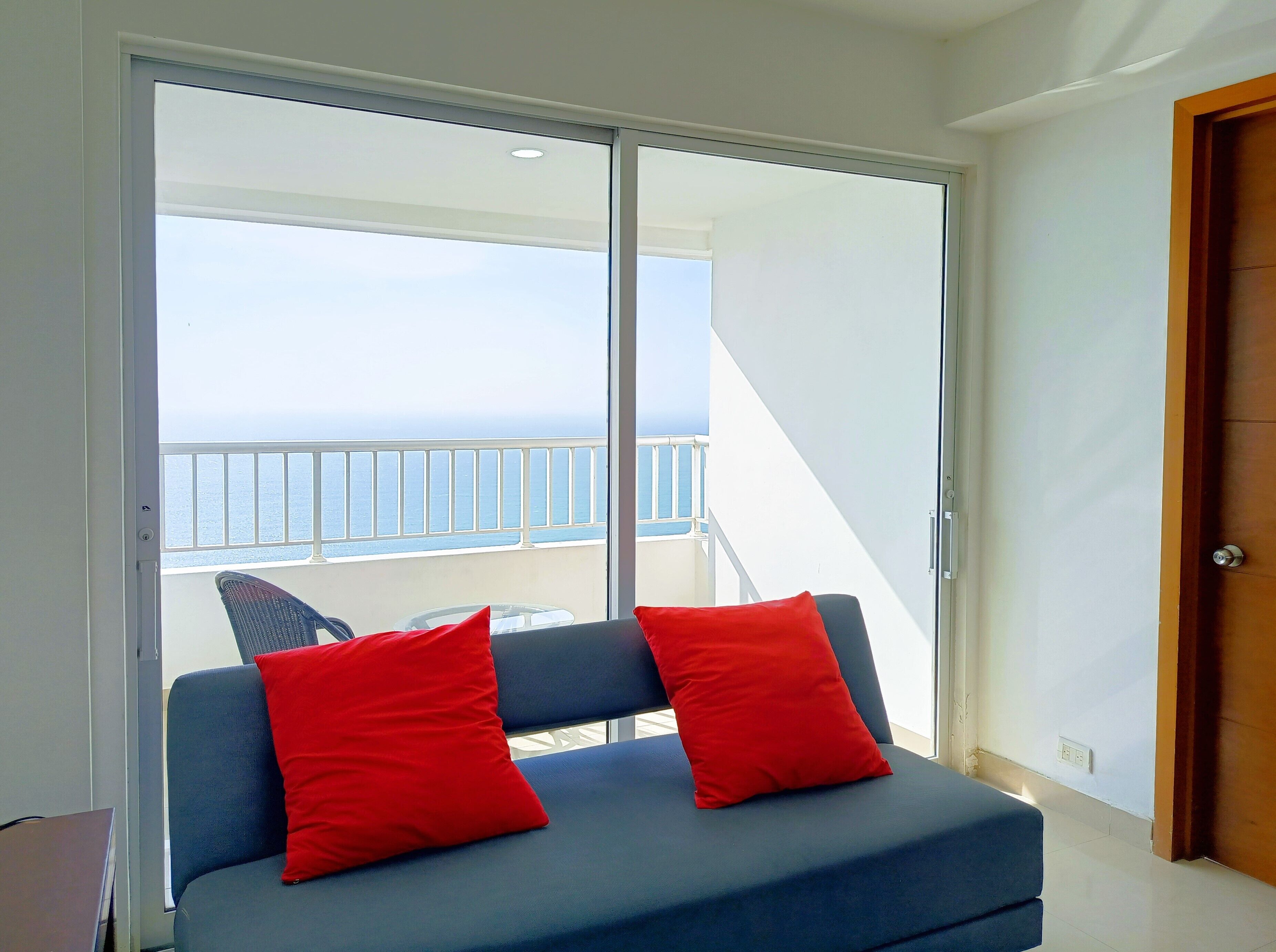 Ein Zimmer mit einem blauen Sofa, zwei roten Kissen und Meerblick durch eine Schiebetür.