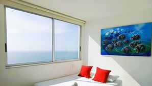 Habitación de hotel con un ventanal grande, una cama con ropa de cama blanca y almohadas rojas, y un cuadro de peces en la pared.