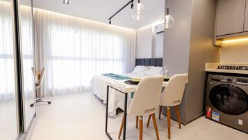 A modern hotel room with a dining area, a bed, a washing machine, and a large window with curtains.