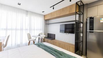 A modern hotel room with a bed, a desk, a chair, a television, and a refrigerator.