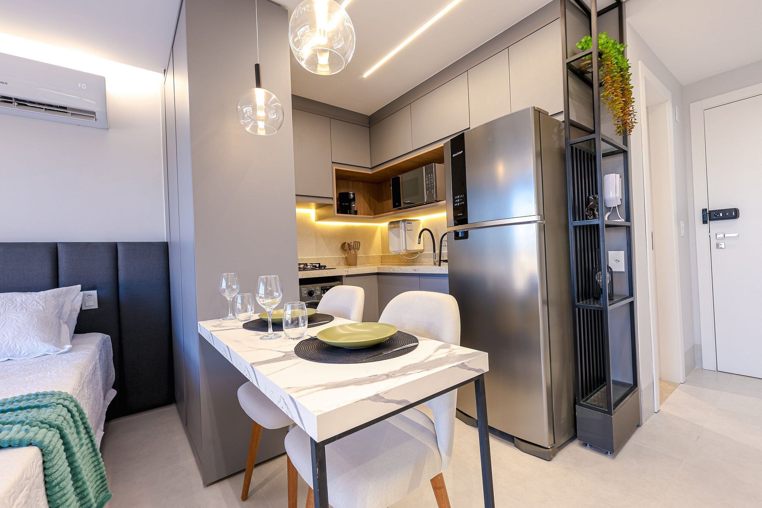 A modern hotel room with a bed, a dining table set with plates and glasses, a kitchen area with a refrigerator and microwave, and a small plant on a shelf.