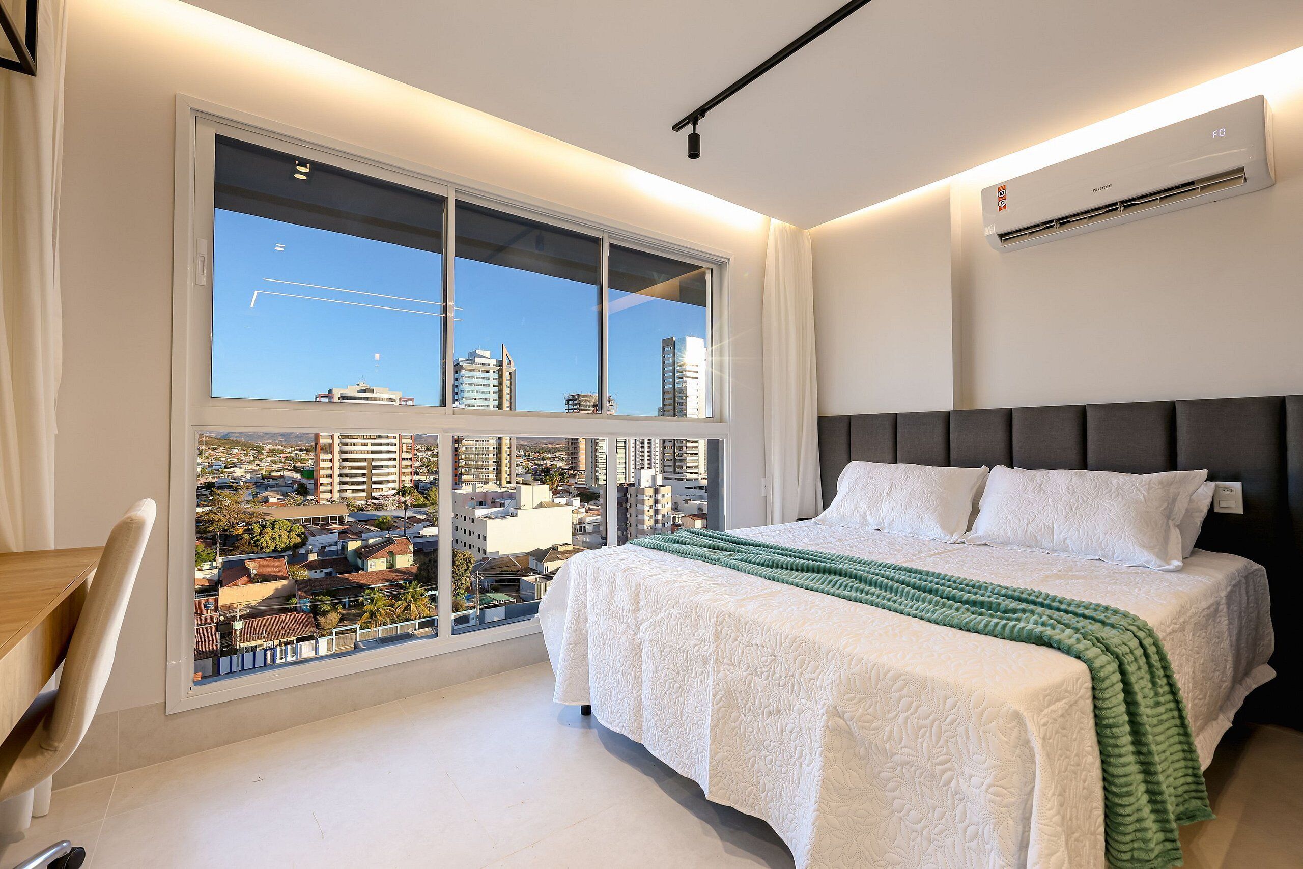A modern bedroom with a large window, a bed with white bedding, and a view of the cityscape.