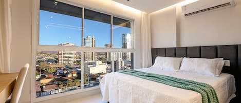A modern bedroom with a large window, a bed with white bedding, and a view of the cityscape.