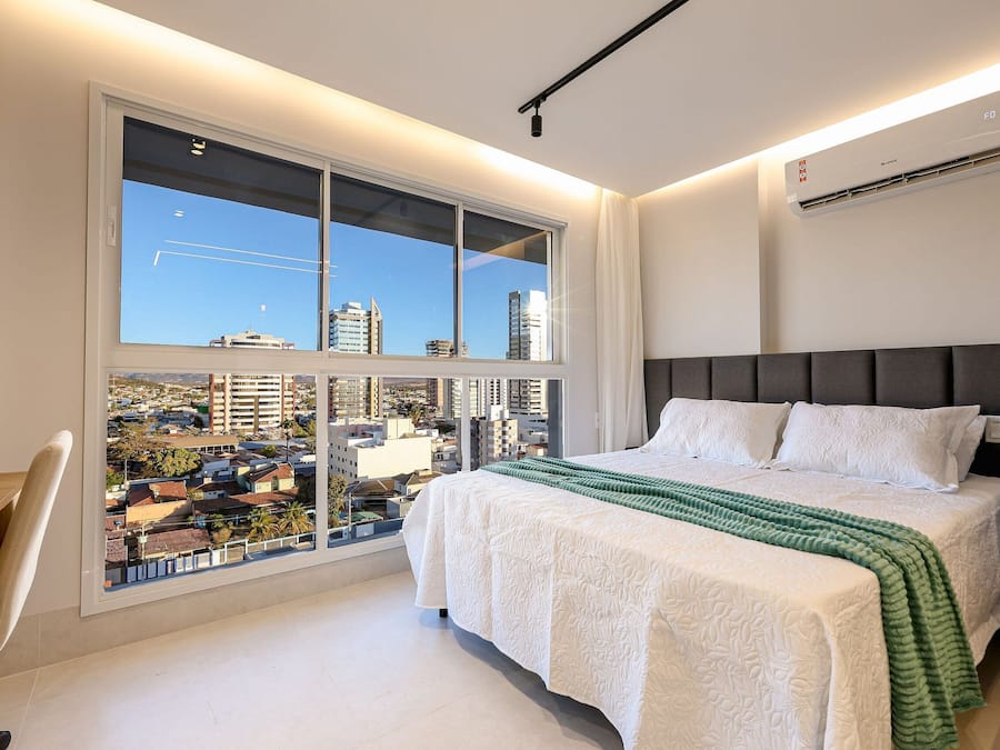 A modern bedroom with a large window, a bed with white bedding, and a view of the cityscape.