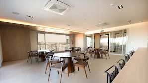A modern dining area with round tables and chairs, large windows offering a city view, and a long counter with chairs. - City House (Barreiras)