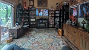 A cozy living room with a fireplace, bookshelves, a television, and a large window overlooking a garden. - 1-bedroom/bath Cozy rustic cottage, off the RCR with AC in lovely Wrinkler, Tx.  (Streetman)