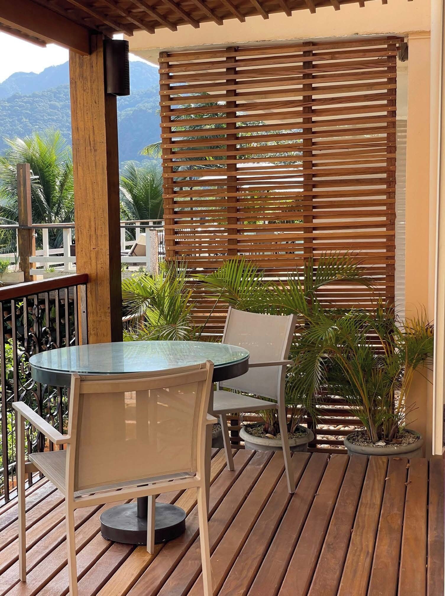 Una terraza de madera con una mesa de vidrio y dos sillas, rodeada de plantas en macetas y una celosía de madera.