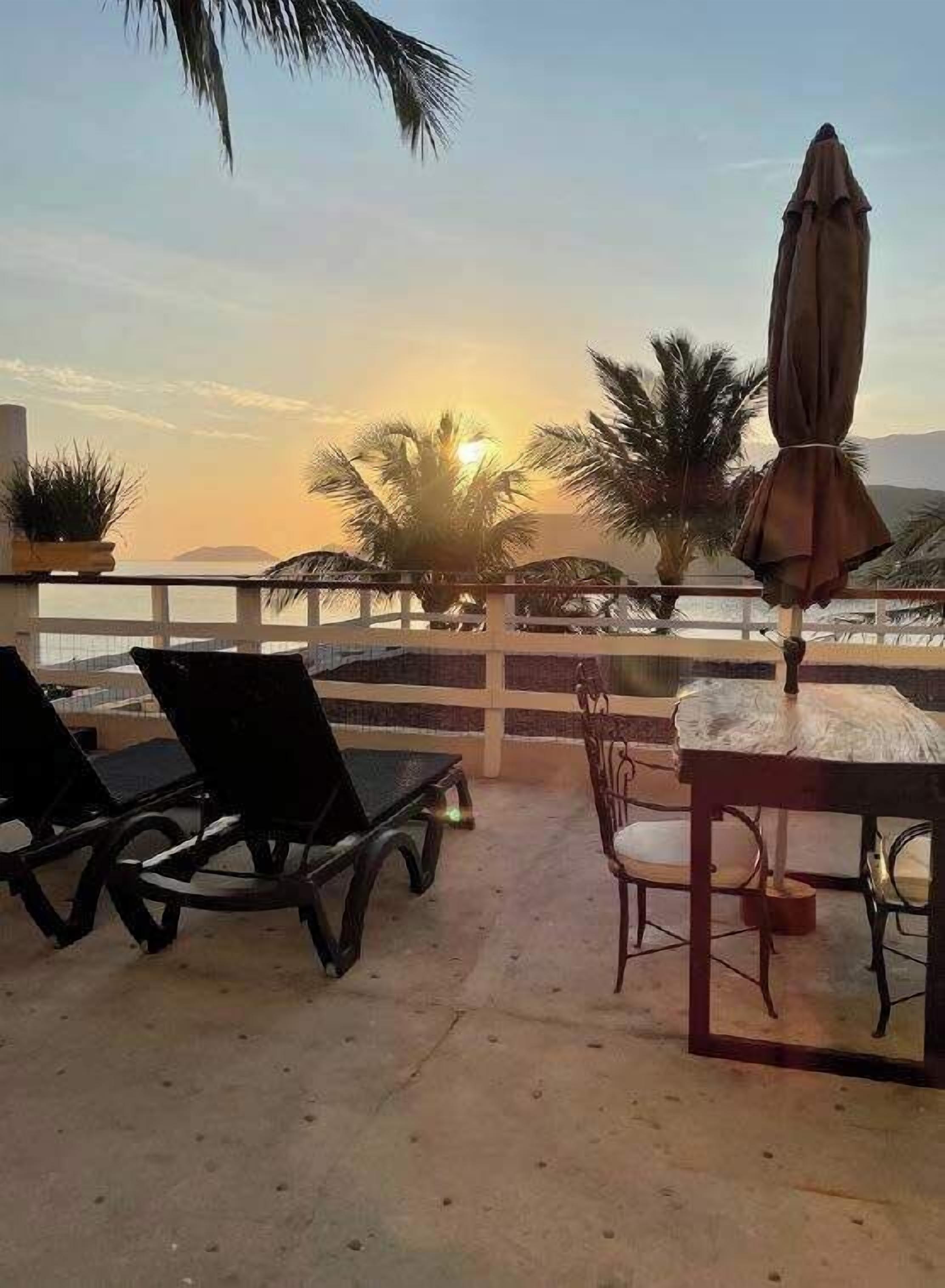 A terrace with lounge chairs, a table, and an umbrella, overlooking palm trees and a sunset.