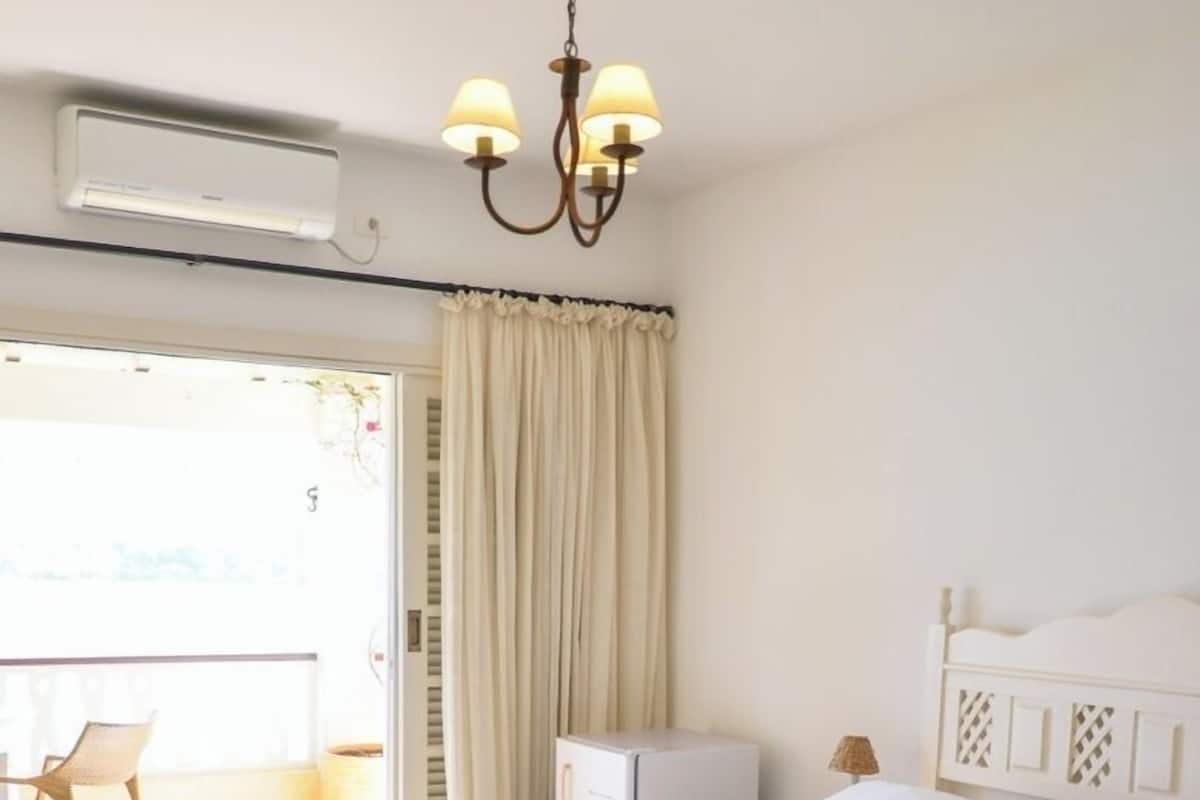 Ένα δωμάτιο ξενοδοχείου με ένα κρεβάτι, ένα μικρό γραφείο, μια καρέκλα, ένα μπαλκόνι και έναν ανεμιστήρα οροφής.