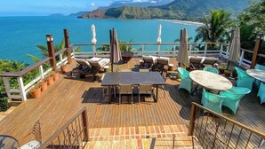 A wooden deck with outdoor seating, including tables and chairs, overlooking a coastal area with a view of the sea and mountains. - Villa Dei Fiori (São Sebastião)
