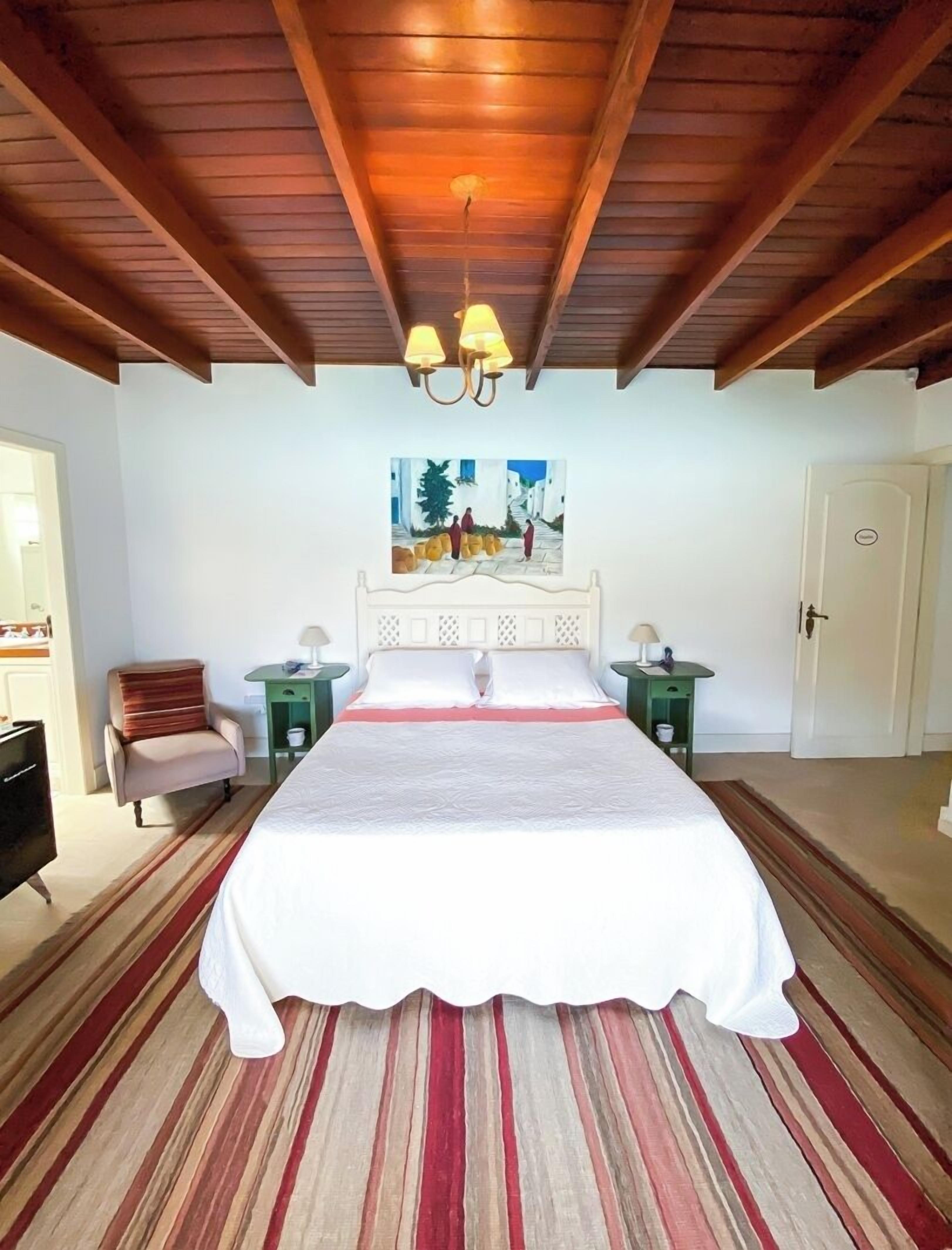 A bedroom with a large bed, wooden ceiling beams, a painting above the bed, and a chair to the left.