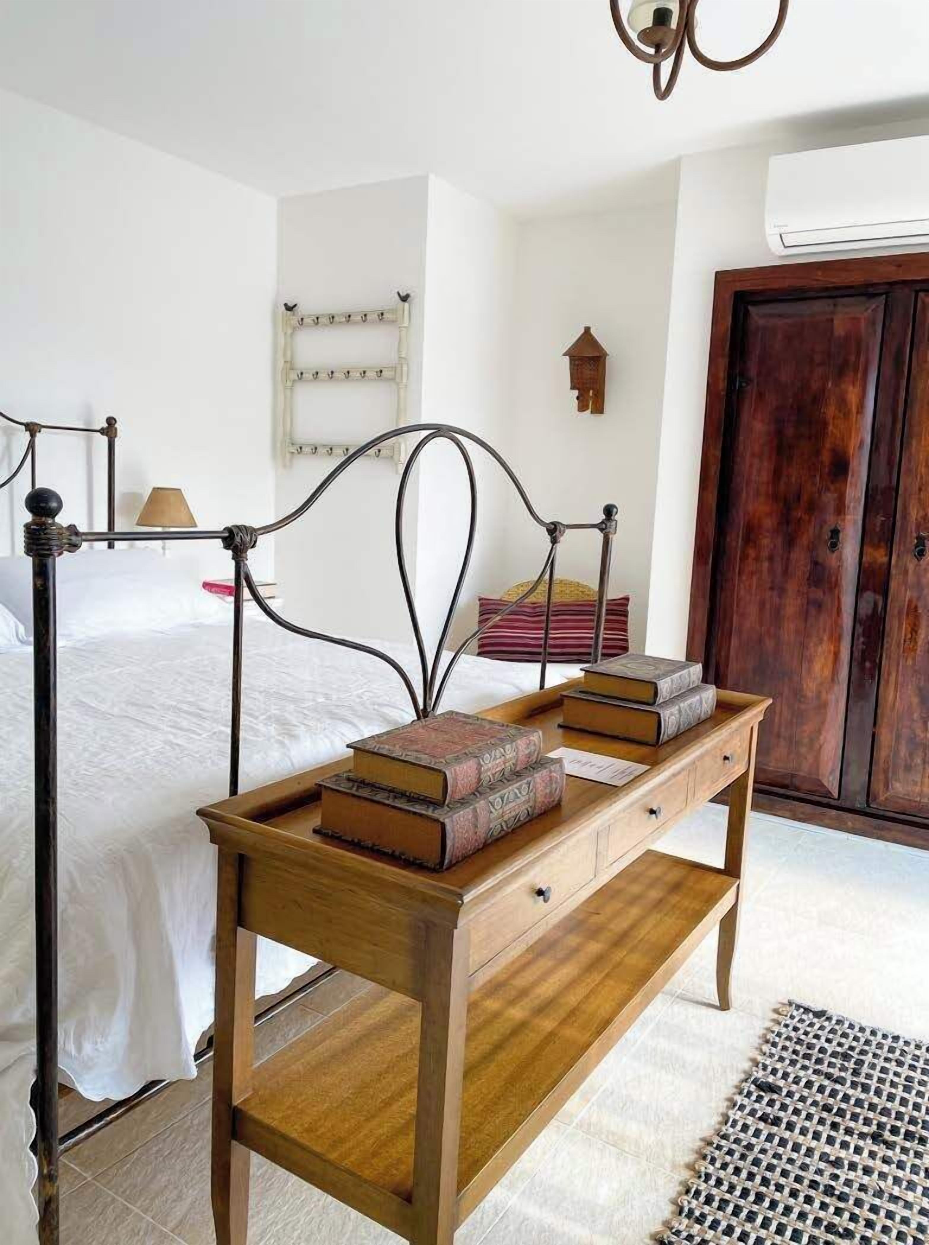Une chambre à coucher avec un lit, une table de chevet en bois sur laquelle sont posés des livres, une lampe, une étagère fixée au mur et une armoire en bois.