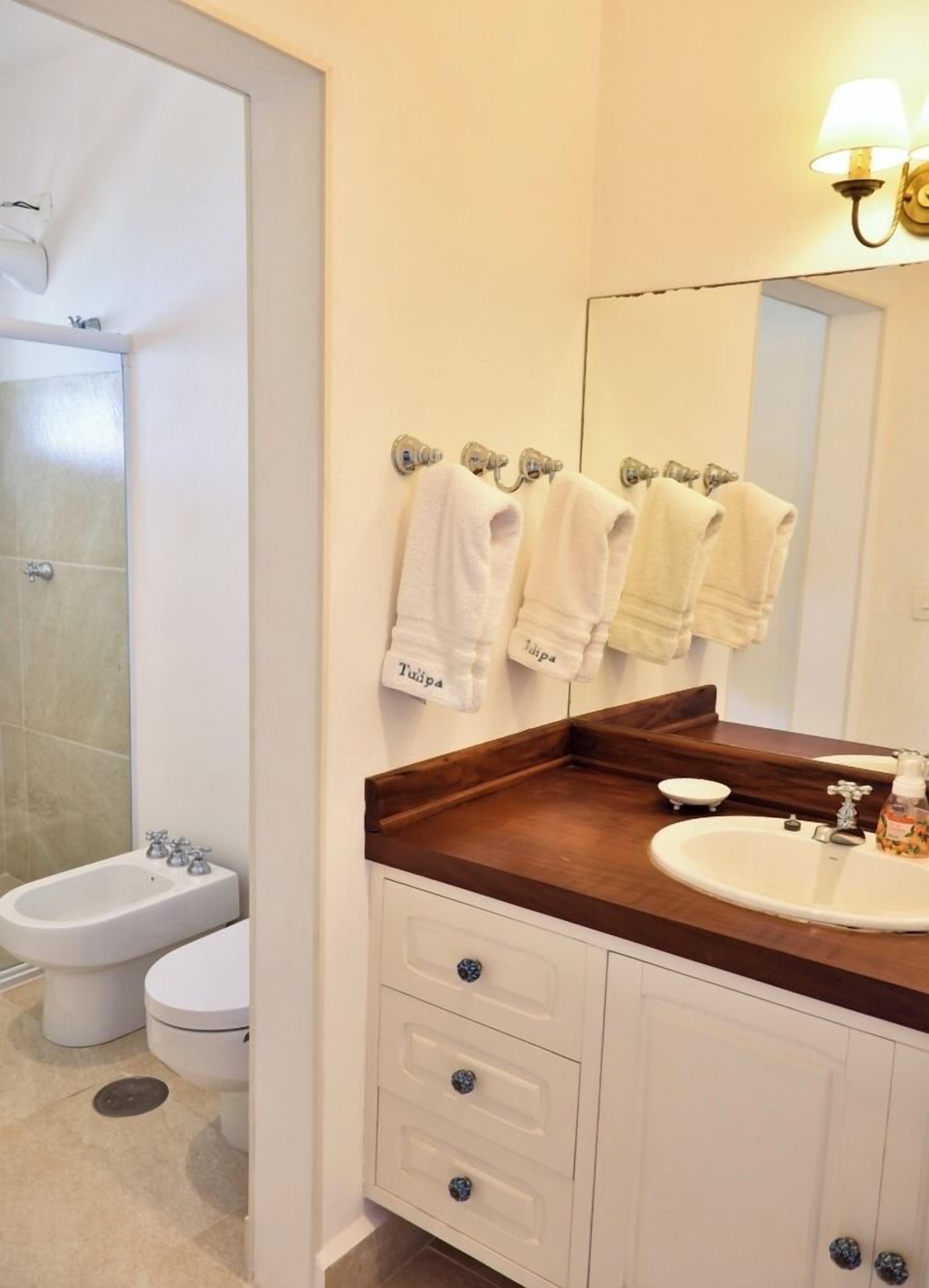 A bathroom with a white sink, wooden countertop, and a mirror above it. There are three towels hanging on a towel rack, and a bidet is visible to the left.