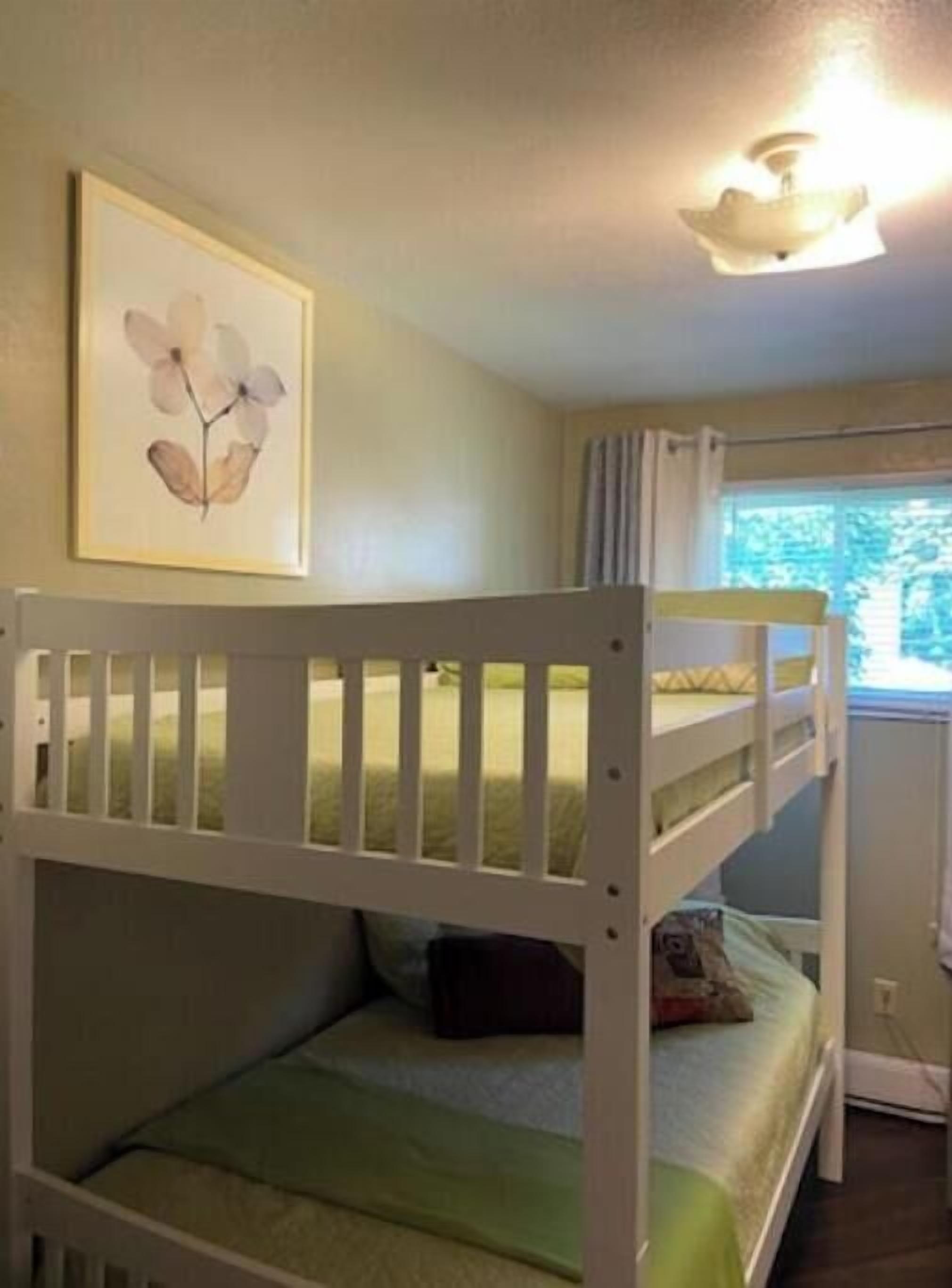 A bunk bed room with a framed floral artwork on the wall, a window with curtains, and a ceiling light fixture.