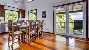 A spacious kitchen with wooden flooring, a dining table with chairs, and large windows offering a view of palm trees.
