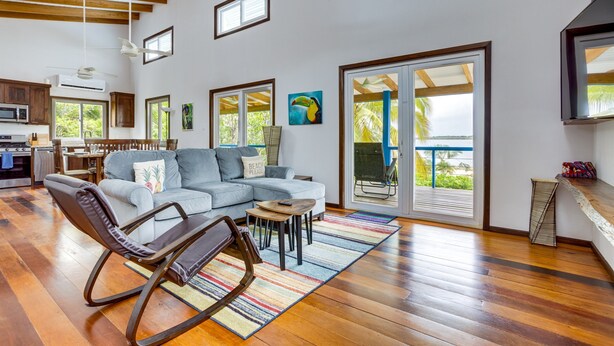 A spacious living area with a gray sectional sofa, a rocking chair, and a small coffee table. There is a large sliding glass door leading to a balcony with a view of palm trees.