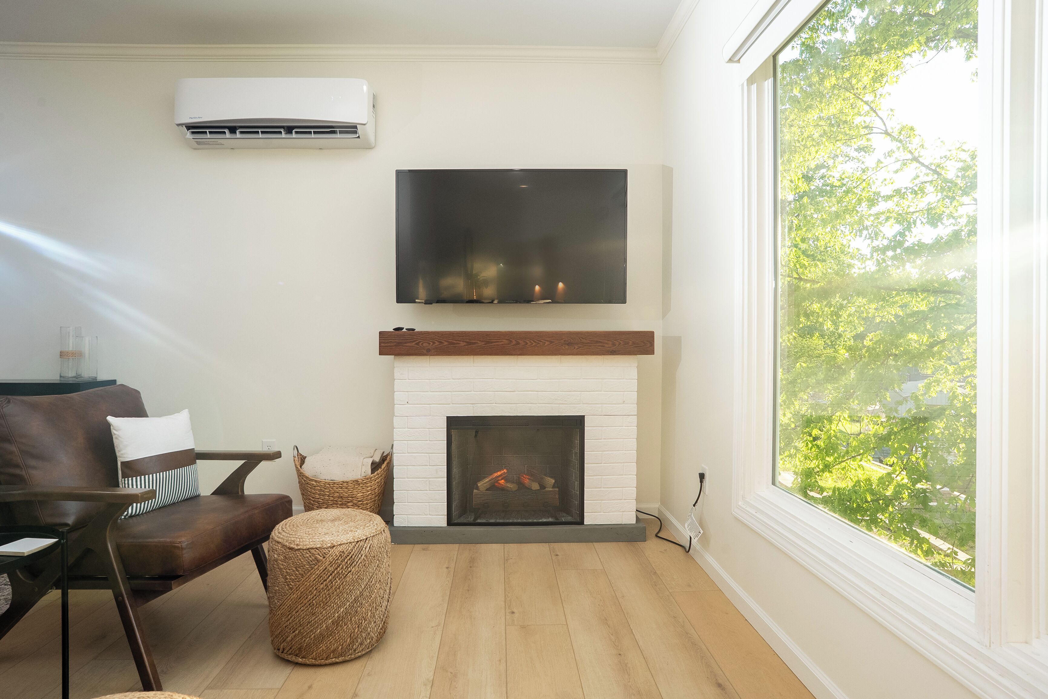 Una sala de estar con chimenea blanca, televisor montado, sofá de cuero marrón y un amplio ventanal con vista a árboles.