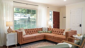 Un salon comprenant un canapé d’angle, une table d’appoint avec une lampe, un fauteuil vert et une vue sur l’extérieur grâce à une grande fenêtre.