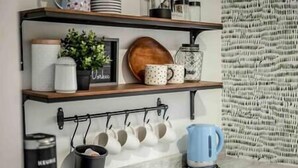 Een keuken met een houten plank waarop mokken, een potplant en een decoratief bord staan. Daaronder een rek met witte mokken, en een blauwe waterkoker op het aanrecht.