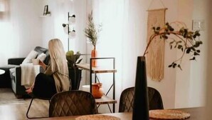 Een moderne woonkamer met een bank, een eettafel en stoelen. Op de tafel staat een plant en erboven een hanglamp.