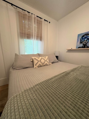 Ein Schlafzimmer mit einem Bett, einem Fenster mit durchsichtigen Vorhängen, einem Bild an der Wand und einem Nachttisch.