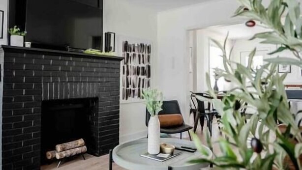 Un salon moderne avec une cheminée en briques noires, un téléviseur à écran plat fixé au-dessus, et une table basse ronde agrémentée d’un vase et de livres.