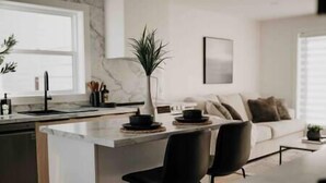 Eine moderne Küche mit Marmor-Spritzschutz, einer zentralen Kochinsel mit Stühlen und einem Essbereich mit Sofa und Kissen.