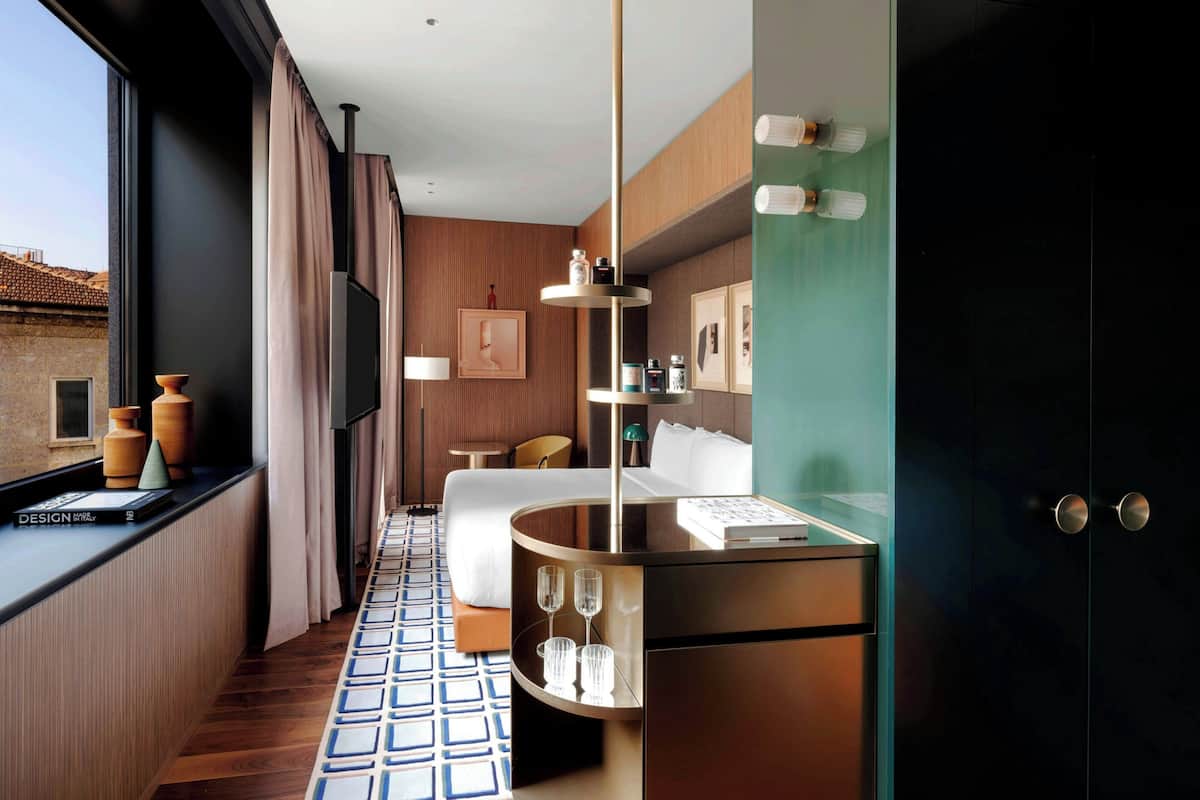 A modern hotel room with a bed, bedside table, and a view of the cityscape.