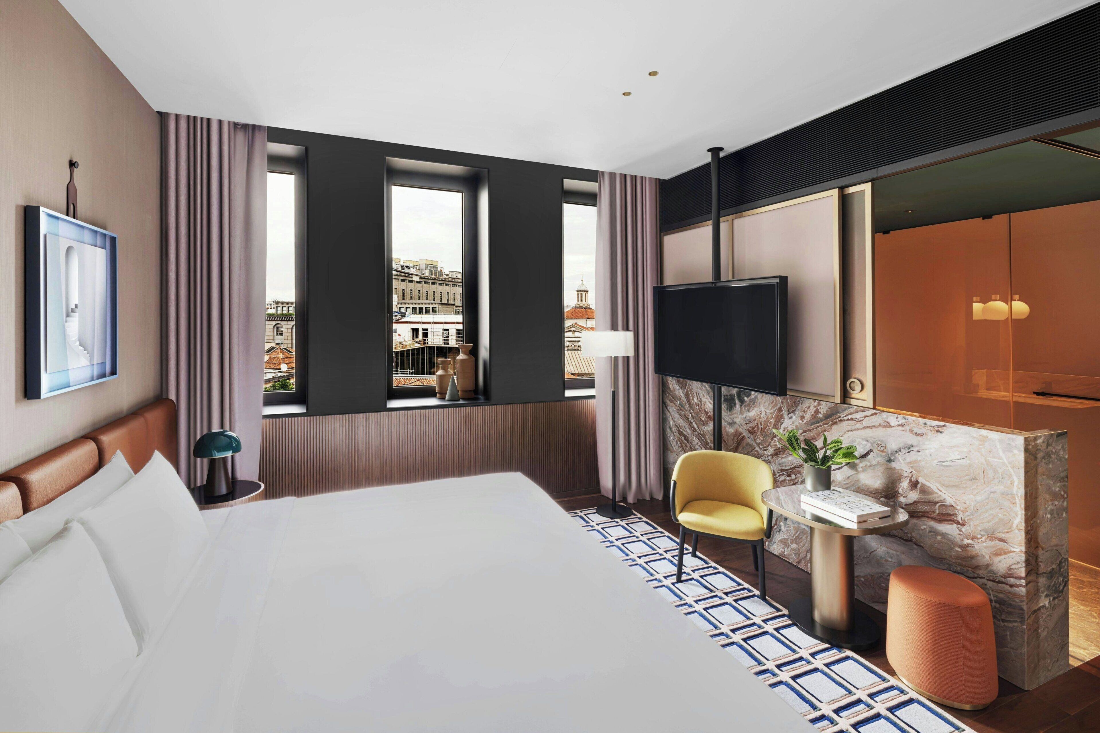 Une chambre d’hôtel moderne avec un grand lit, un bureau agrémenté d’une plante, une chaise et une vue sur la ville grâce à de grandes fenêtres.
