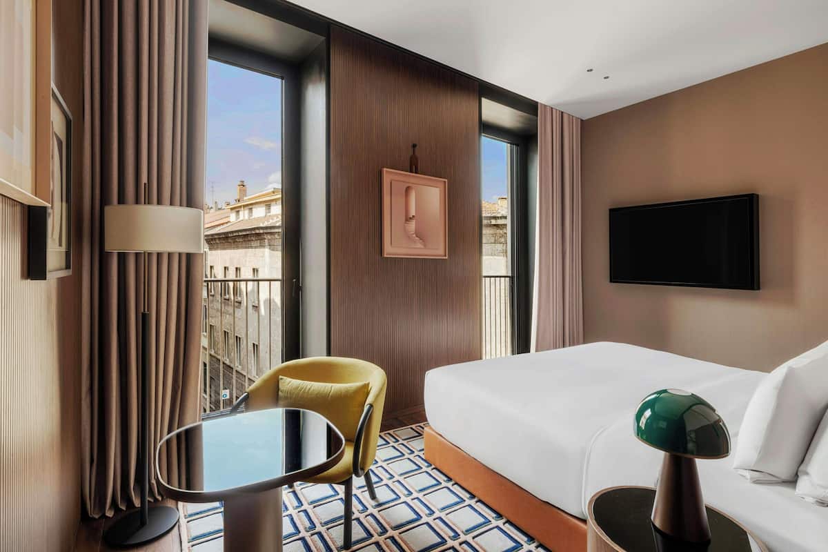 A hotel room with a bed, a desk, a chair, a TV, and a window with a city view.