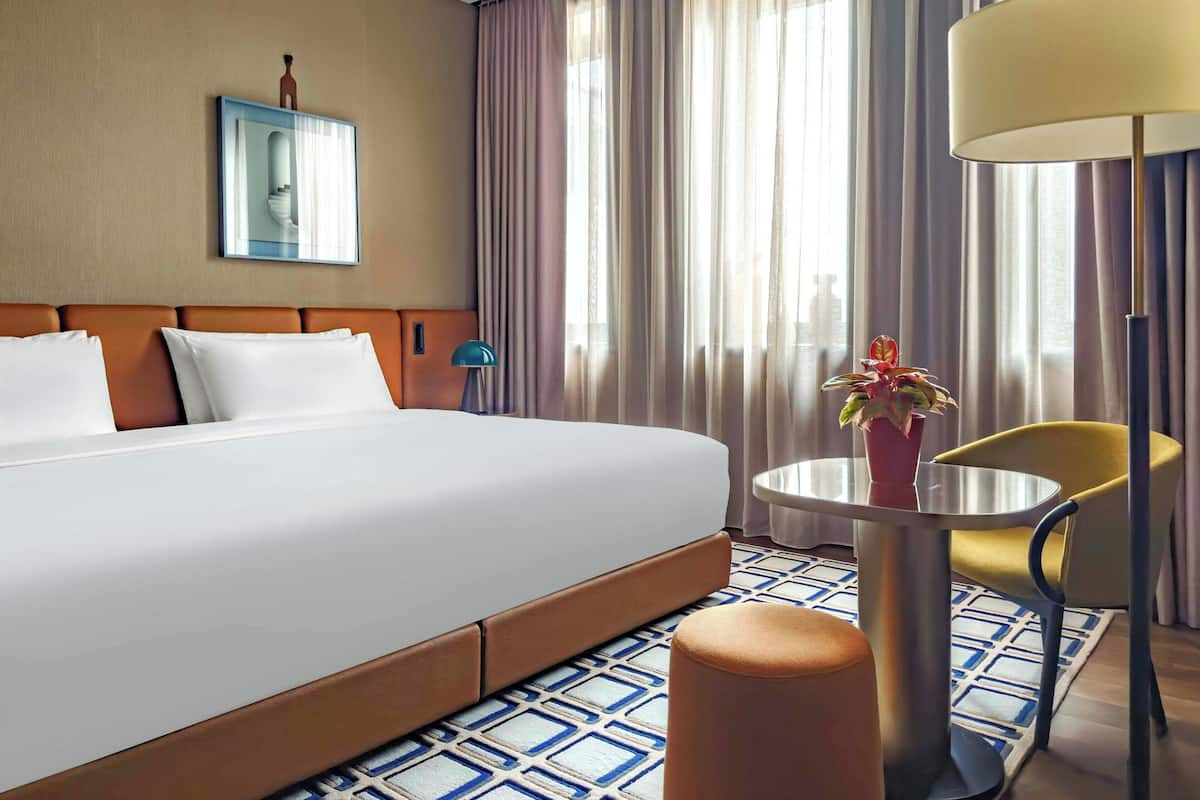 A hotel room with a large bed, a round table, a chair, a lamp, and a potted plant.