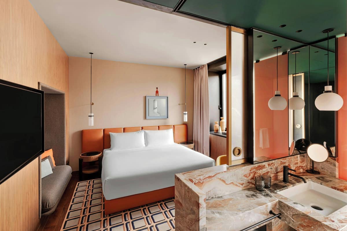 A modern hotel room with a large bed, a TV, a seating area, a bathroom with a marble sink, and a large mirror.